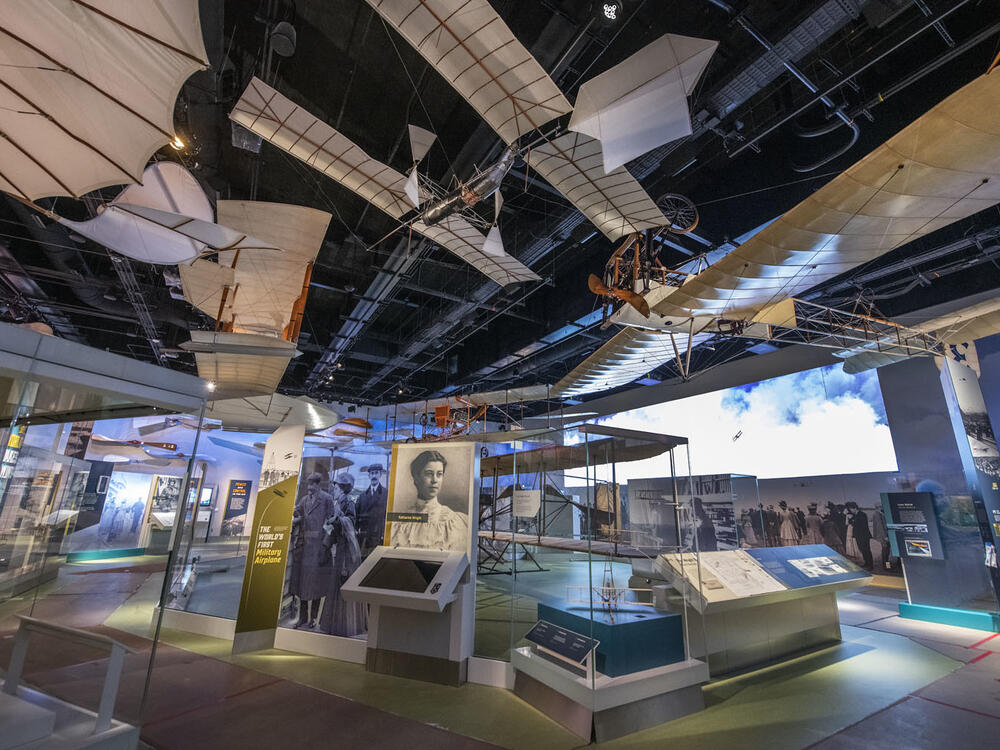 Several beige biplanes hang from the ceiling over exhibitry. 