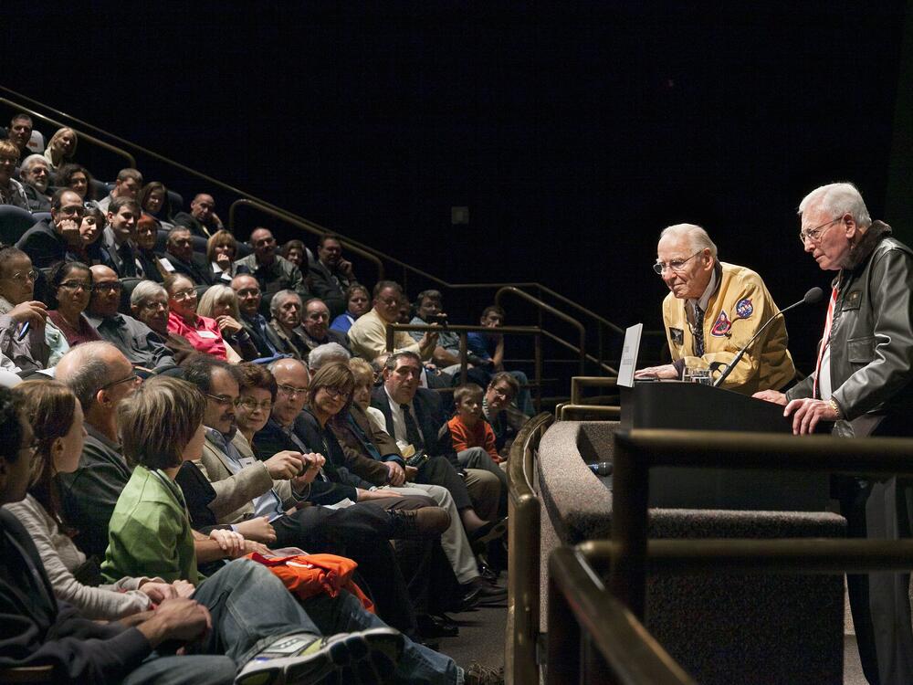 A speaker, an older white man, leans over the podium to talk to a young person. 