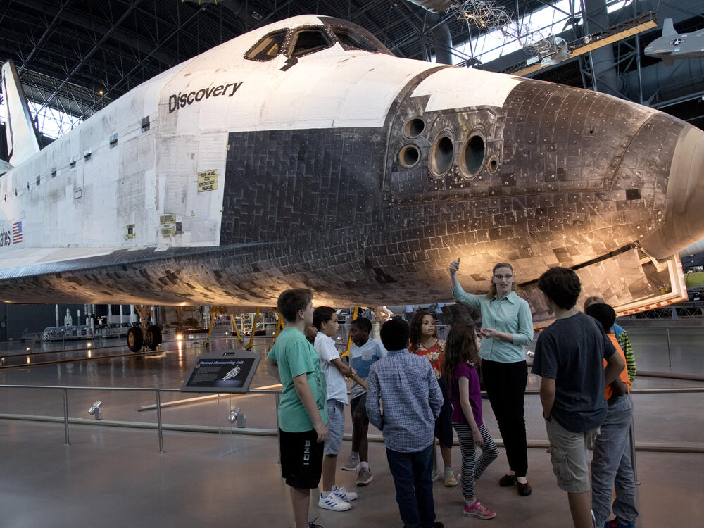 A group of school children and an educator stand in front of the Space Shuttle Discovery. 