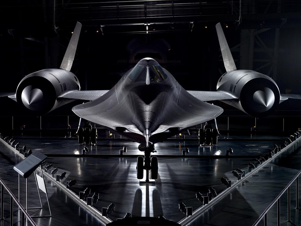A black spy plane stands in an all black air hanger, lit from the light from above. The curvy form of the plane--bulging at two engines on either wing and curving up toward the cockpit--give the sense of an almost science fiction aircraft (although it's very real).