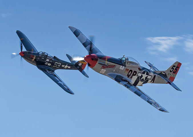 The dark blue P-51D Man O War plane races through the sky alongside Pretty Polly, a P-63C plane with a grey fuselage and blue-striped wings.