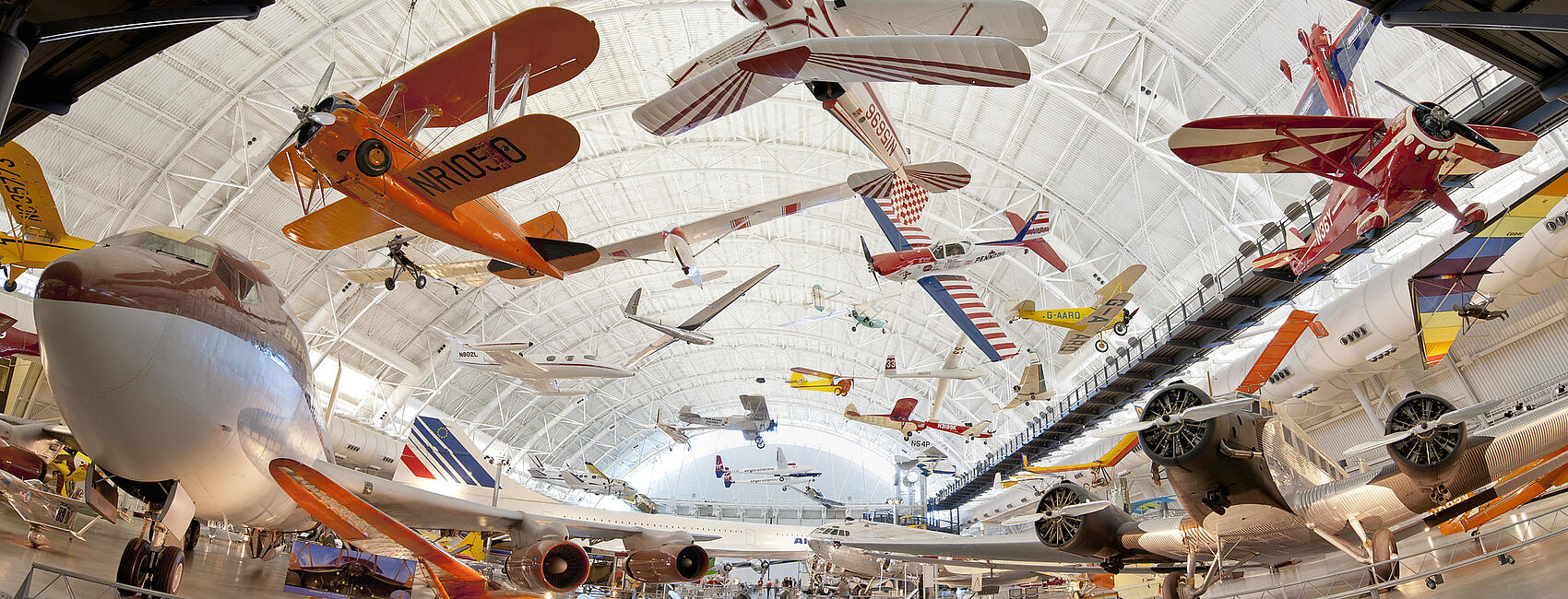 Dozens of various aircraft of all type hang suspended from the ceiling in a large hangar. More aircraft dot the ground.